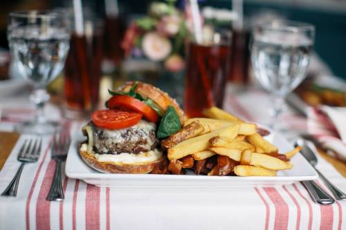 A plate with a cheeseburger topped with tomatoes and pickles, served with golden fries and drinks in the background.