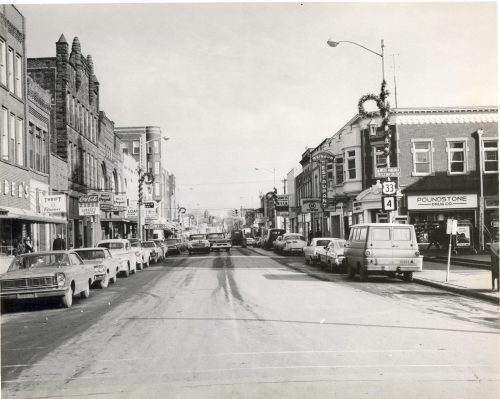 Black and white photo of a busy street lined with vintage cars and storefronts, decorated for the holidays.