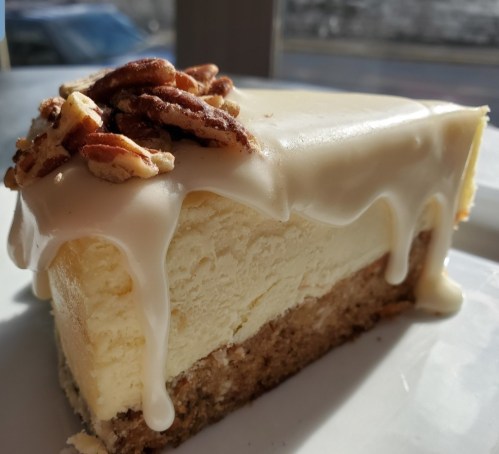 A slice of creamy cheesecake topped with drizzled icing and pecans, resting on a white plate.