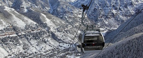 A ski gondola ascends a snowy mountain, surrounded by trees and a valley below.