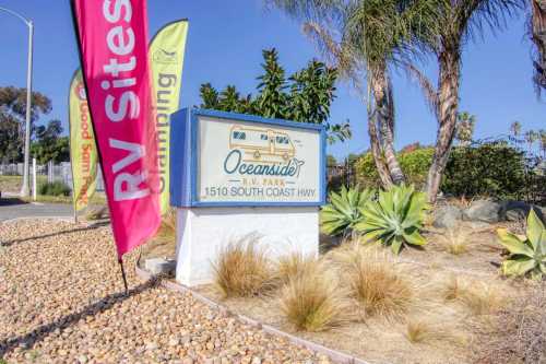 Sign for Oceanside RV Park at 1510 South Coast Highway, surrounded by plants and colorful flags.