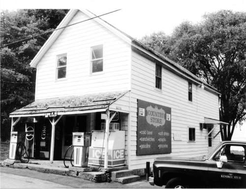 A vintage black-and-white photo of a country store with gas pumps and a sign advertising ice and corn.