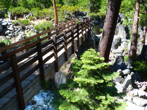 A wooden bridge spans a rocky area, surrounded by trees and greenery in a natural setting.