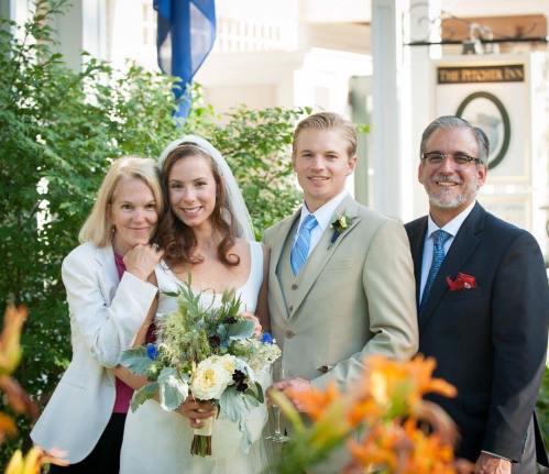 A bride and groom pose with two adults, smiling, surrounded by greenery and flowers at a wedding venue.