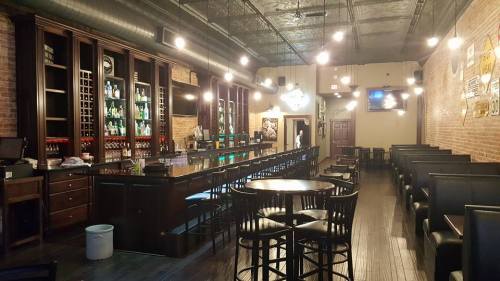 A cozy bar interior with a long counter, wooden tables, and brick walls, illuminated by hanging lights.