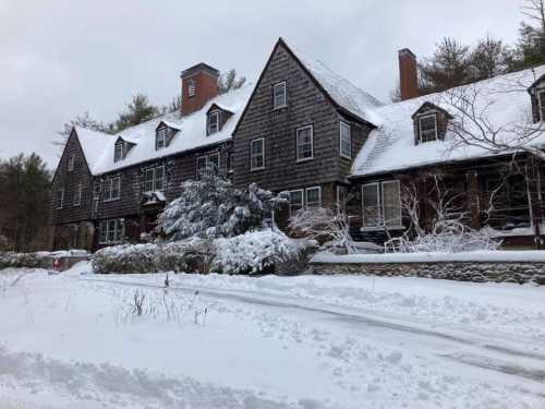 A large, snow-covered house with a steep roof and multiple chimneys, surrounded by trees and a snowy landscape.