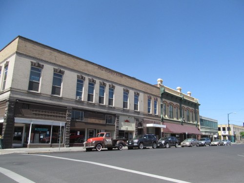 Historic buildings line a sunny street, featuring shops and parked cars under a clear blue sky.