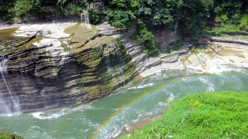 A scenic view of a waterfall cascading over layered rocks, with a vibrant rainbow arching over a flowing river below.
