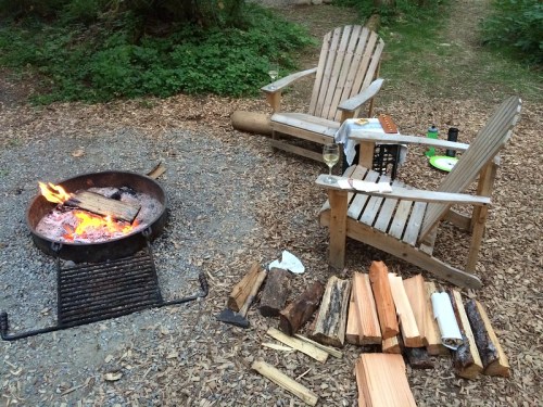 A cozy campfire scene with two wooden chairs, firewood stacked nearby, and a glowing fire in a fire pit.