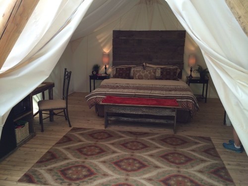 Cozy tent interior featuring a bed, wooden decor, lamps, and a patterned rug on a wooden floor.