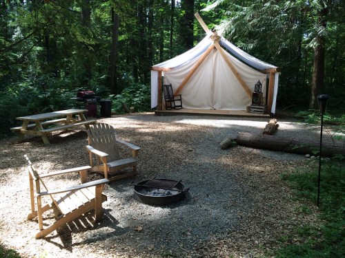 A serene forest setting with a tent, picnic table, and wooden chairs around a fire pit on a gravel area.