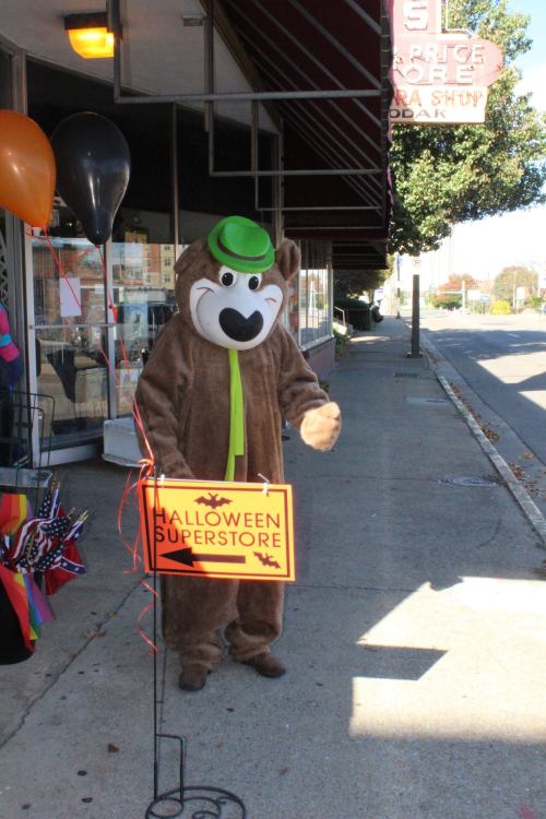 A person in a bear costume with a green hat stands outside a store, holding a sign that says "Halloween Superstore."
