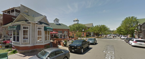 A charming street scene featuring shops, outdoor seating, and parked cars under a clear blue sky.