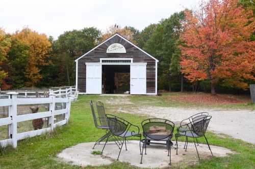 A rustic barn with a sign, surrounded by colorful autumn trees and a fire pit with chairs in the foreground.