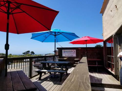 Outdoor seating area with red and blue umbrellas, overlooking a scenic view. Sign for "Queenie's Roadhouse Cafe" visible.