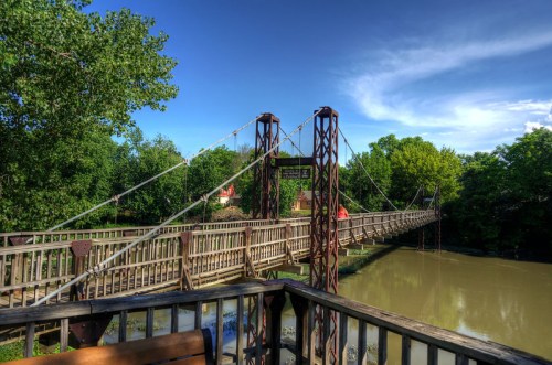 A suspension bridge spans a river, surrounded by lush green trees and a clear blue sky.