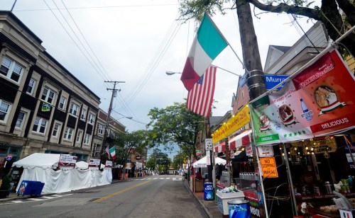 A street lined with food stalls and flags, featuring Italian and American decorations, under a cloudy sky.
