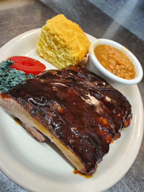 A plate featuring glazed barbecue ribs, cornbread, baked beans, a slice of tomato, and a garnish of kale.