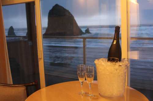 A bottle of champagne in an ice bucket with two glasses, overlooking a coastal view through a window.