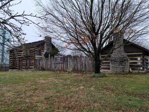 Historic log cabin structures with a wooden fence, set in a grassy area under a cloudy sky.
