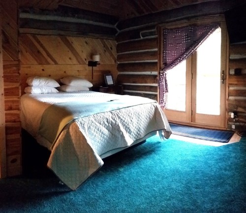 Cozy log cabin bedroom with a bed, wooden walls, and a door leading to an outdoor area. Soft lighting and carpeted floor.