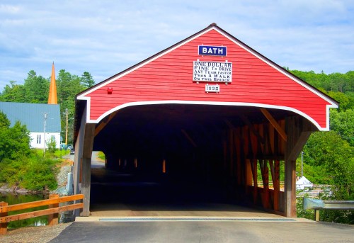 A red covered bridge with a sign reading "Bath" and "One Dollar Fine to Pass" surrounded by greenery and a blue sky.