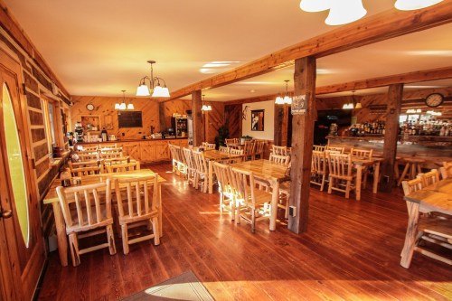 Cozy restaurant interior with wooden tables and chairs, warm lighting, and a bar area in the background.