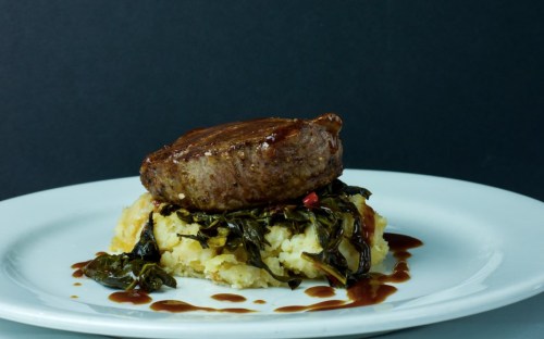 A plate featuring a tender beef patty atop creamy mashed potatoes and sautéed greens, drizzled with sauce.