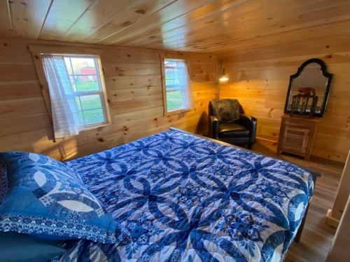 Cozy bedroom with a wooden interior, featuring a blue quilted bed, a chair, and natural light from windows.