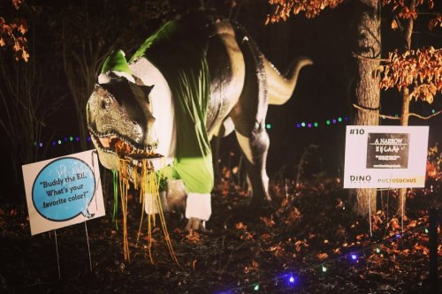 A dinosaur statue dressed in a green outfit, surrounded by festive lights and signs in a wooded area.