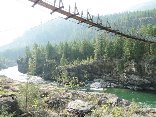 A wooden suspension bridge spans a river surrounded by lush green trees and rocky cliffs in a misty landscape.