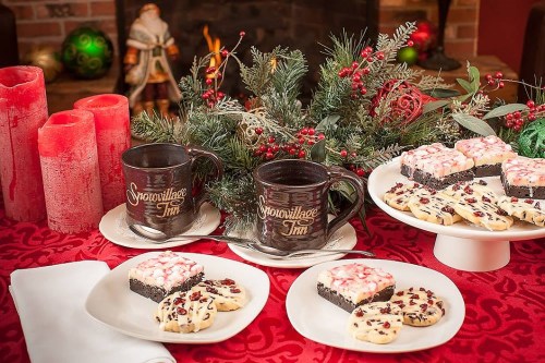A festive table setting with two mugs, holiday treats, and candles, surrounded by greenery and Christmas decorations.