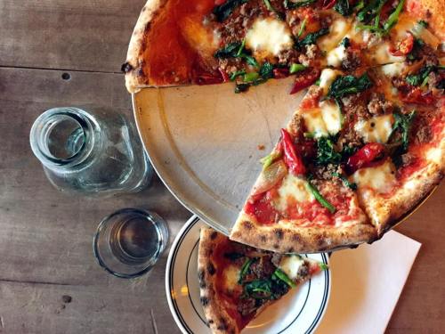 A pizza topped with cheese, greens, and red peppers, with a slice removed, on a wooden table next to glassware.