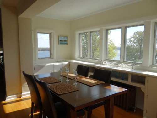 Bright dining area with a wooden table, chairs, and large windows overlooking a scenic lake view.