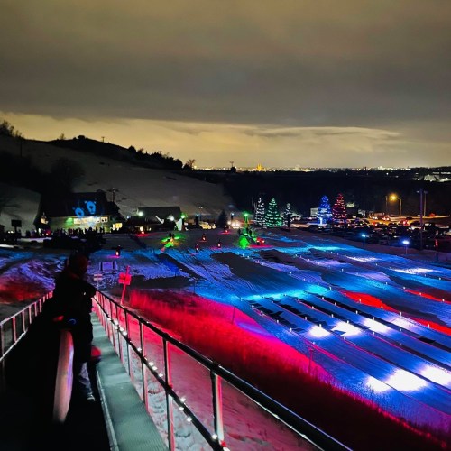 A snowy landscape at night, illuminated by colorful lights, with trees and a slope for winter activities in the background.