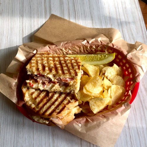A grilled sandwich with crispy chips and a pickle slice, served in a red basket lined with brown paper.