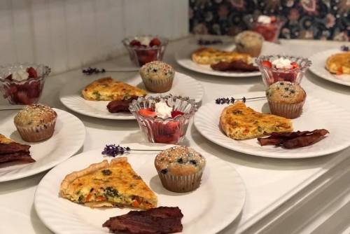 A table set with plates of quiche, muffins, bacon, and fruit cups, garnished with flowers.