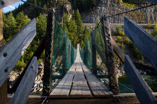 A wooden suspension bridge stretches over a river, surrounded by lush greenery and rocky cliffs.
