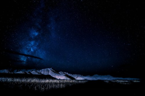A starry night sky over snow-capped mountains, with a dark landscape and a hint of grass in the foreground.
