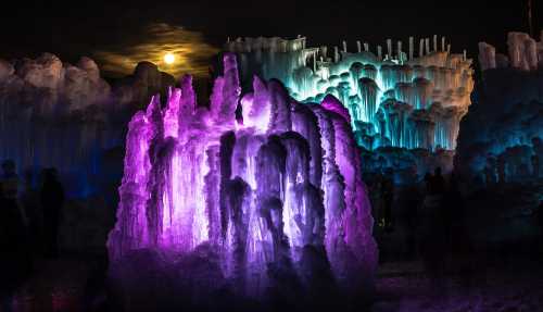 Colorful ice formations illuminated by purple and blue lights, with a full moon in the background.