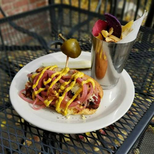 A hot dog topped with mustard and pickled onions, served with a side of colorful chips in a metal cup.