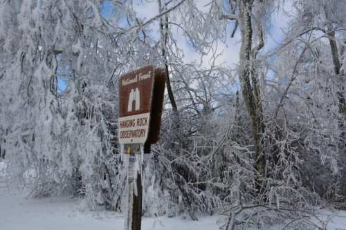 A sign for Hanging Rock Observatory surrounded by trees covered in ice and snow, under a partly cloudy sky.