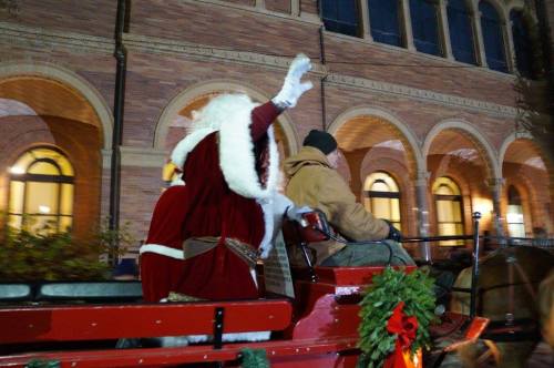 A person in a Santa costume waves from a festive horse-drawn carriage, with holiday decorations visible.