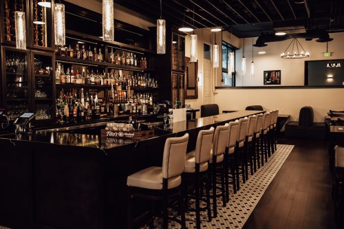 A stylish bar with a long counter, high stools, and shelves filled with various bottles in a modern setting.