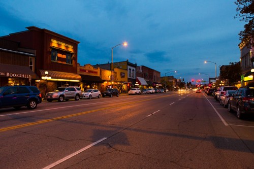 A quiet street at dusk, lined with shops and parked cars, illuminated by streetlights and a blue sky.
