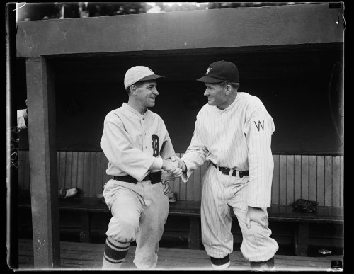 Two baseball players shake hands in a dugout, smiling and wearing their team uniforms.