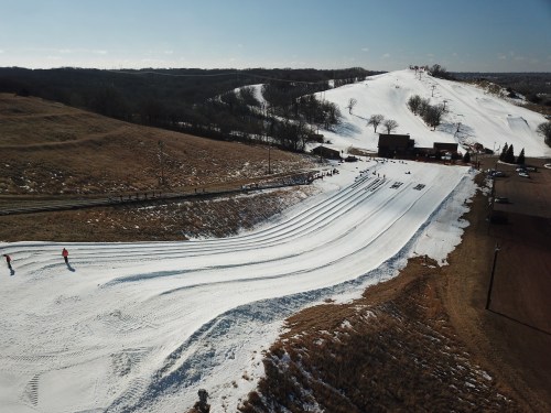 A snowy hill with ski tracks, people skiing, and a lodge in the background under a clear blue sky.