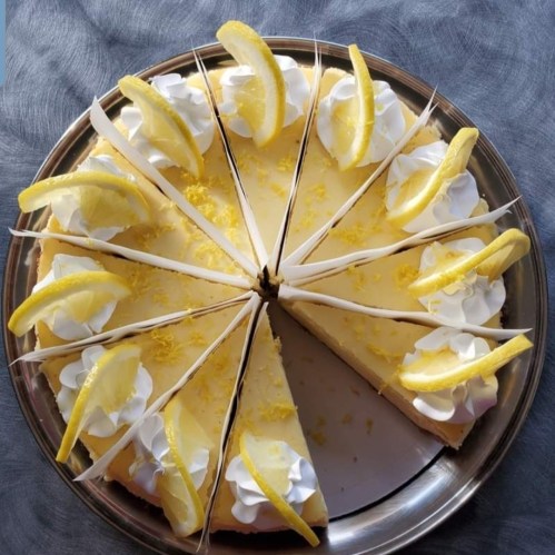 A round lemon cheesecake topped with whipped cream and lemon slices, sliced into equal pieces on a silver platter.