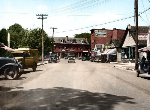 A colorized vintage street scene featuring classic cars, storefronts, and power lines in a small town.