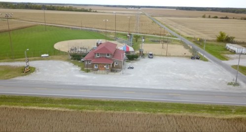 Aerial view of a small building near a sports field, surrounded by farmland and a road.